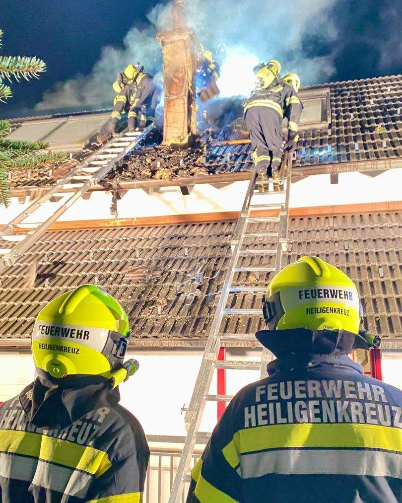 Feuerwehrleute sind auf dem Dach eines Hauses mit aufsteigendem Rauch. Sie benutzen Leitern, um auf das Dach zu gelangen. Sie tragen gelbe Helme mit den Worten 'Feuerwehr' und 'Heilgenkreuz' darauf.
