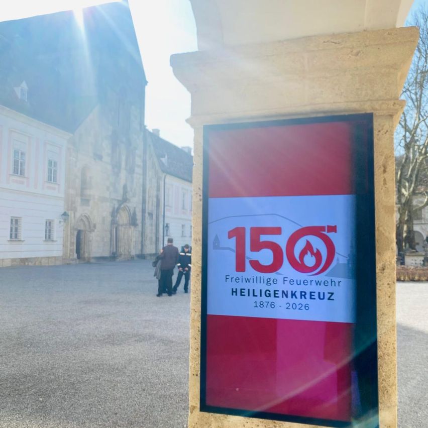Ein Schild vor einem Gebäude mit dem Text '150 Freiwillige Feuerwehr Heiligenkreuz 1876-2026'. Im Hintergrund laufen zwei Personen.