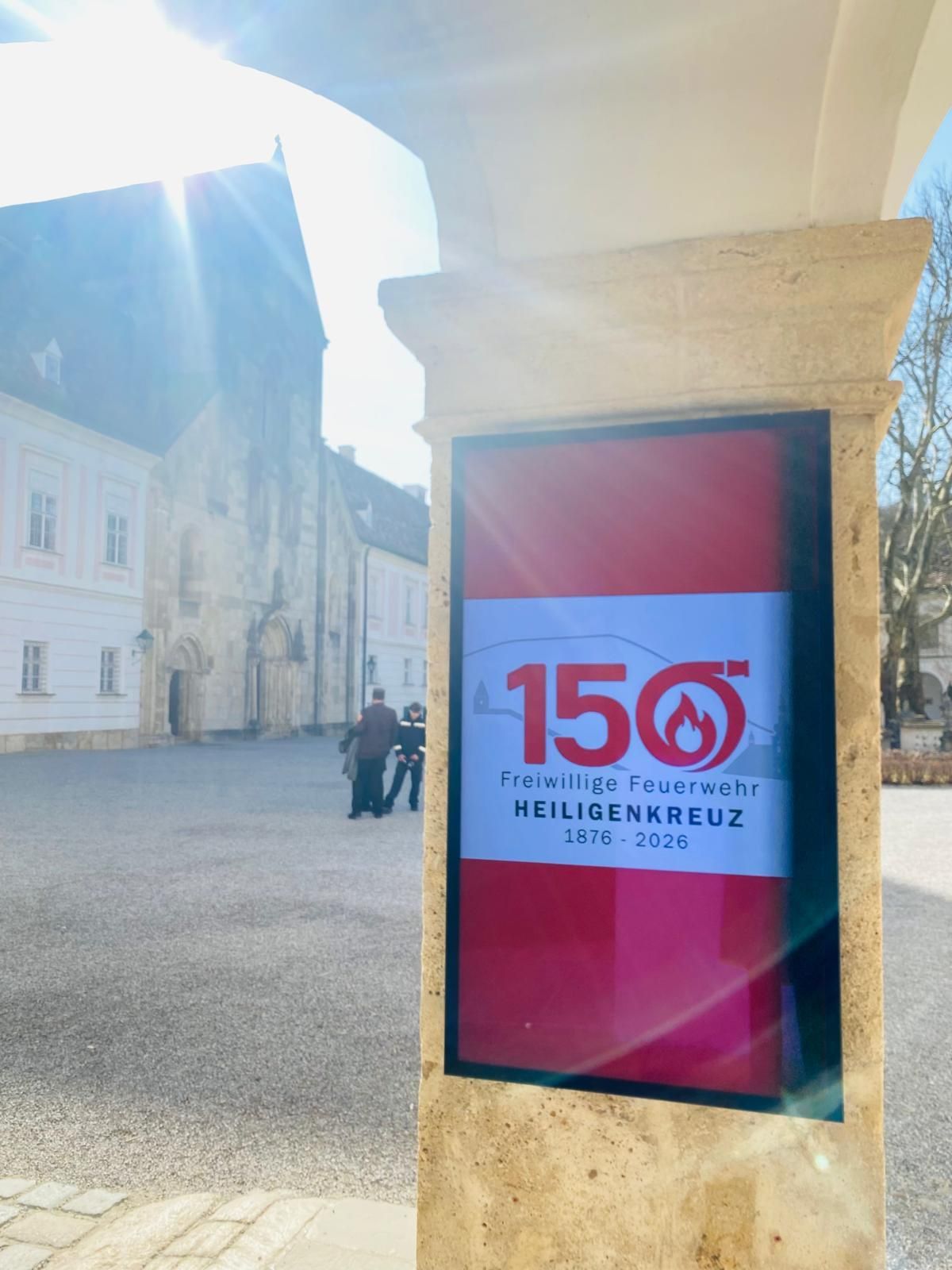 Ein Schild vor einem Gebäude mit dem Text '150 Freiwillige Feuerwehr Heiligenkreuz 1876-2026'. Im Hintergrund laufen zwei Personen.