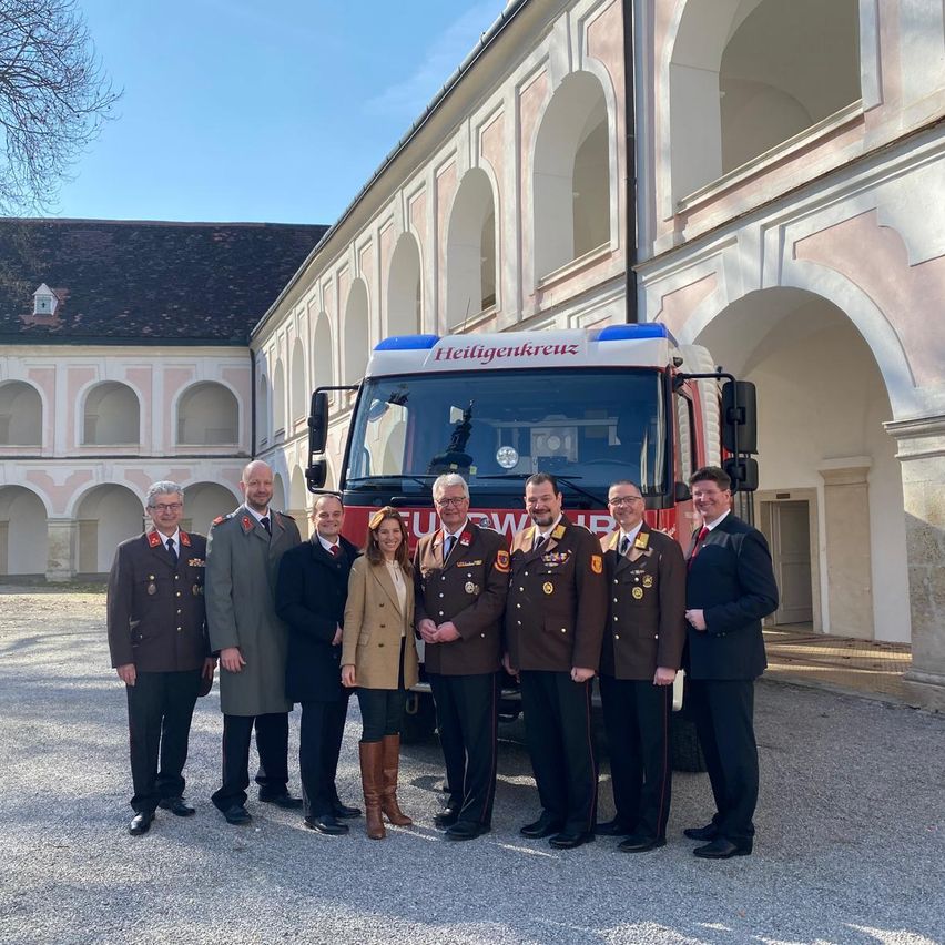 Eine Gruppe uniformierter Personen, darunter Männer und Frauen, steht vor einem roten Feuerwehrauto mit der Aufschrift 'Heilgenkreuz' in einer formellen Umgebung.