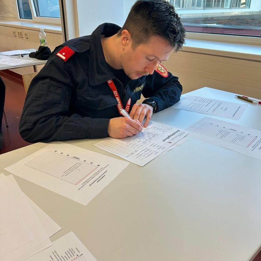 Ein Mann in Uniform schreibt auf einem Blatt Papier an einem Tisch. Er hat einen roten Ausweis mit einem weißen Etikett und der Aufschrift 'PRINZ'. Es gibt mehrere Papiere auf dem Tisch.