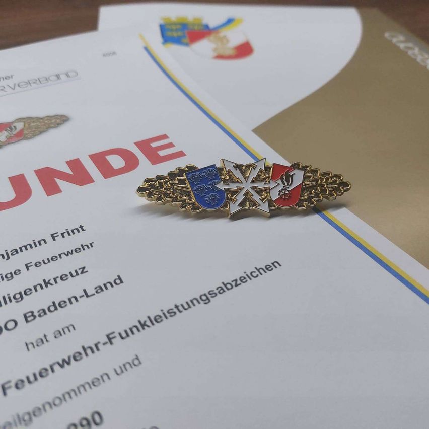 Eine Abzeichen mit einem blauen Schild und rotem Kreuz liegt auf einem Zertifikat, wobei das Wort 'UNDE' in Rot auf dem Papier geschrieben ist. Auf dem Papier steht 'Feuerwehr-Funkleistungsabzeichen'.