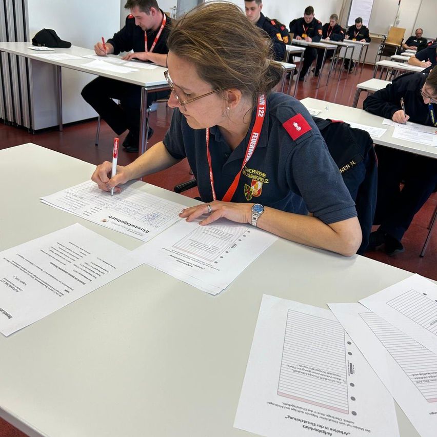 Eine Frau in Uniform schreibt auf einem Stück Papier an einem Schreibtisch. Andere Personen sitzen im Hintergrund an Tischen und schreiben ebenfalls.