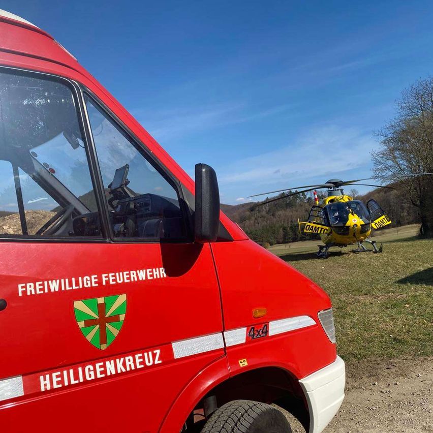 Ein rotes Fahrzeug mit der Aufschrift Freiwillige Feuerwehr Heiligenkreuz ist geparkt. Dahinter steht ein gelber Hubschrauber mit der Aufschrift DAMTC auf dem Rasen.