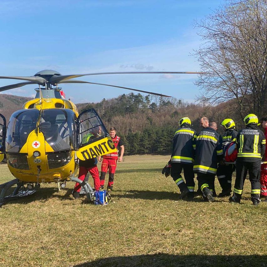Ein gelber Hubschrauber mit DAMTC auf der Seite geparkt auf einem Rasenfeld. Mehrere Feuerwehrleute stehen darum herum, möglicherweise bereit für einen Notfall.