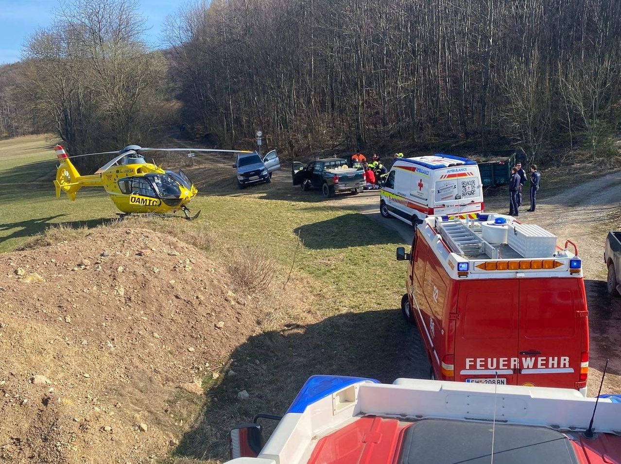 Ein Unfallort mit Rettungsfahrzeugen auf einem Grasfeld. Ein gelber Hubschrauber und ein Krankenwagen sind geparkt. Einsatzkräfte kümmern sich um eine verletzte Person.