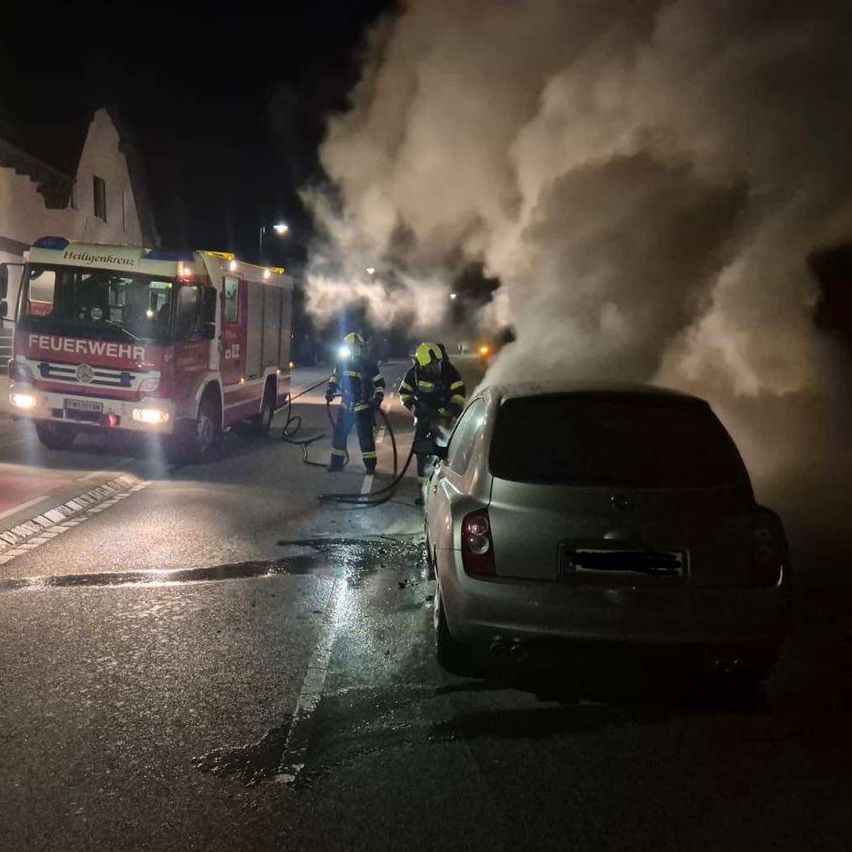 Ein Feuerwehrwagen mit Feuerwehrleuten, die Wasser auf ein rauchendes Auto in der Nacht sprühen.