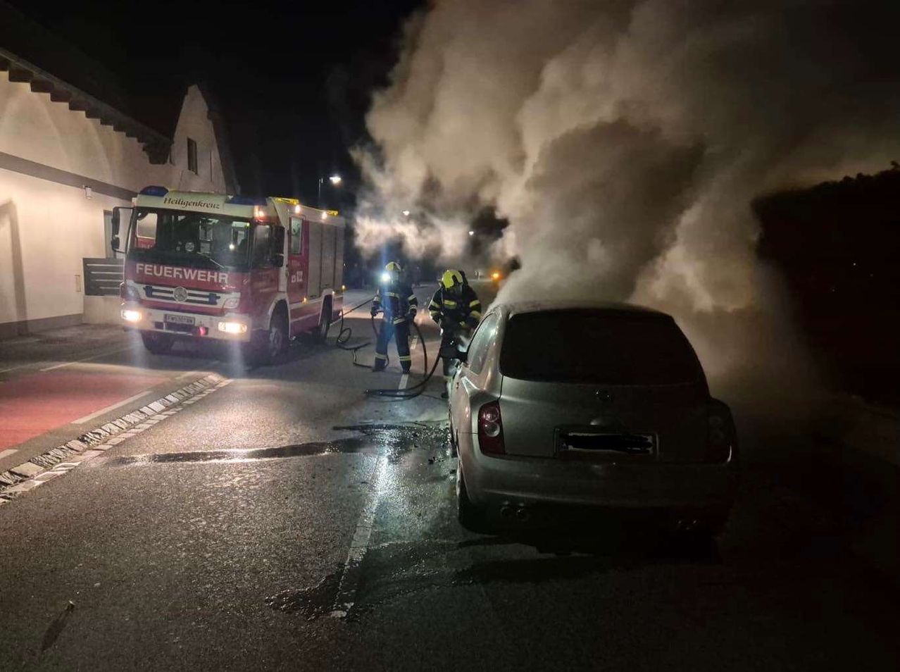 Ein Feuerwehrwagen mit Feuerwehrleuten, die Wasser auf ein rauchendes Auto in der Nacht sprühen.