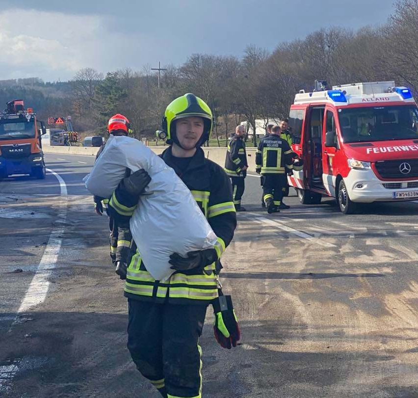 Ein Feuerwehrmann trägt einen weißen Sack auf der Straße, in der Nähe anderer Feuerwehrleute und eines roten Mercedes-Benz-Ambulanzes im Hintergrund.