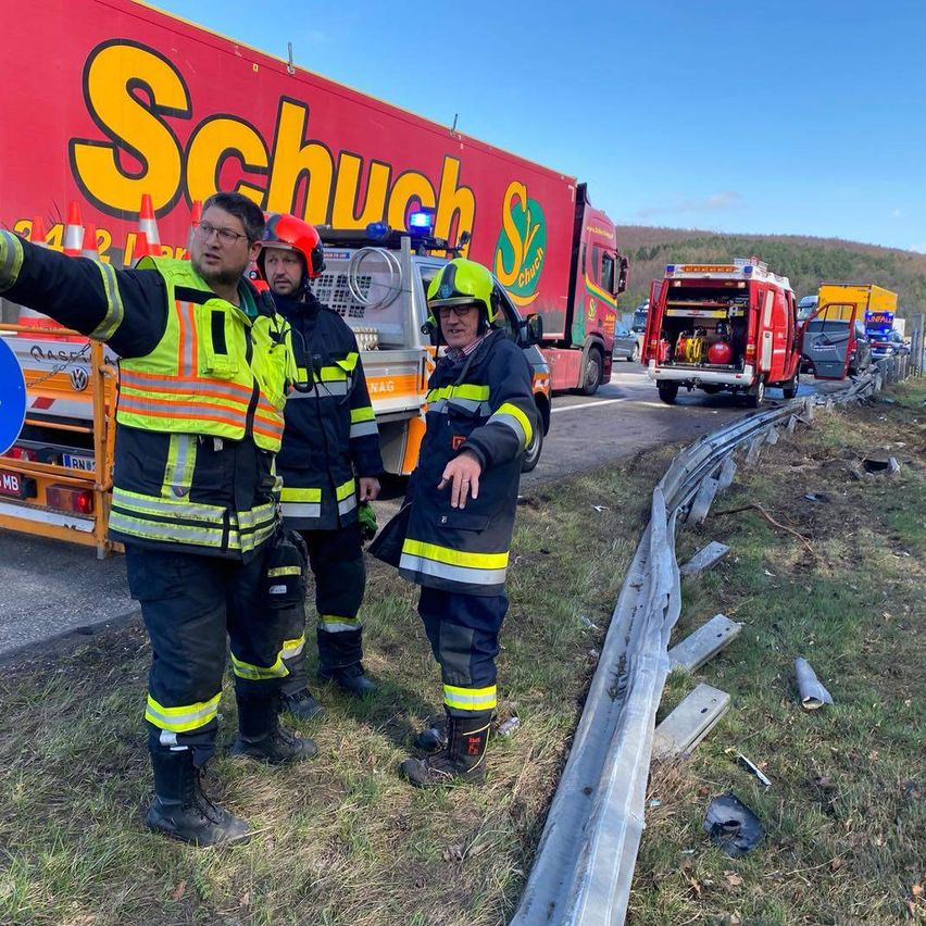 Feuerwehrleute stehen vor einem Unfallort mit Notfallfahrzeugen in der Nähe. Ein Feuerwehrmann zeigt, während ein anderer zusieht. Die Szene umfasst eine beschädigte Leitplanke und Verkehrskegel.