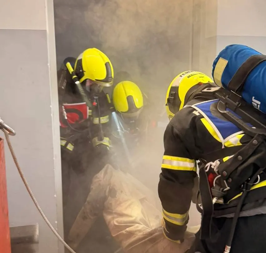 Feuerwehrleute in gelben Helmen und Schutzausrüstung retten jemanden aus einem rauchigen Raum, einer hält ein Seil.