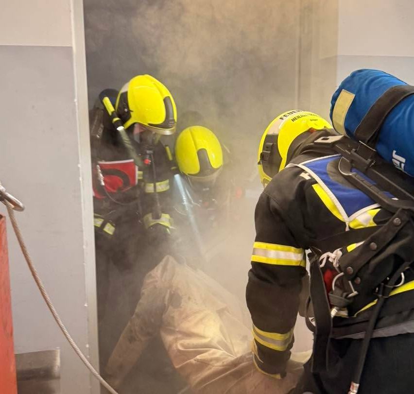Feuerwehrleute in gelben Helmen und Schutzausrüstung retten jemanden aus einem rauchigen Raum, einer hält ein Seil.