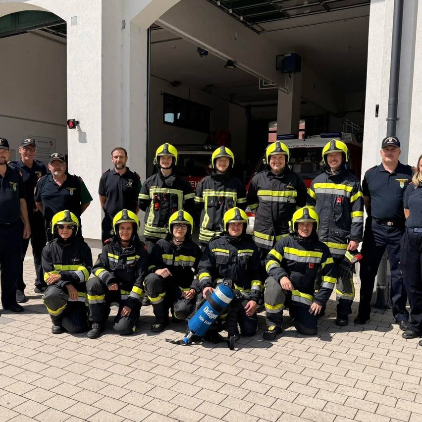 Eine Gruppe von Feuerwehrleuten in Uniform, einige kniend und andere stehend, posiert für ein Foto vor einem Feuerwehrhaus.