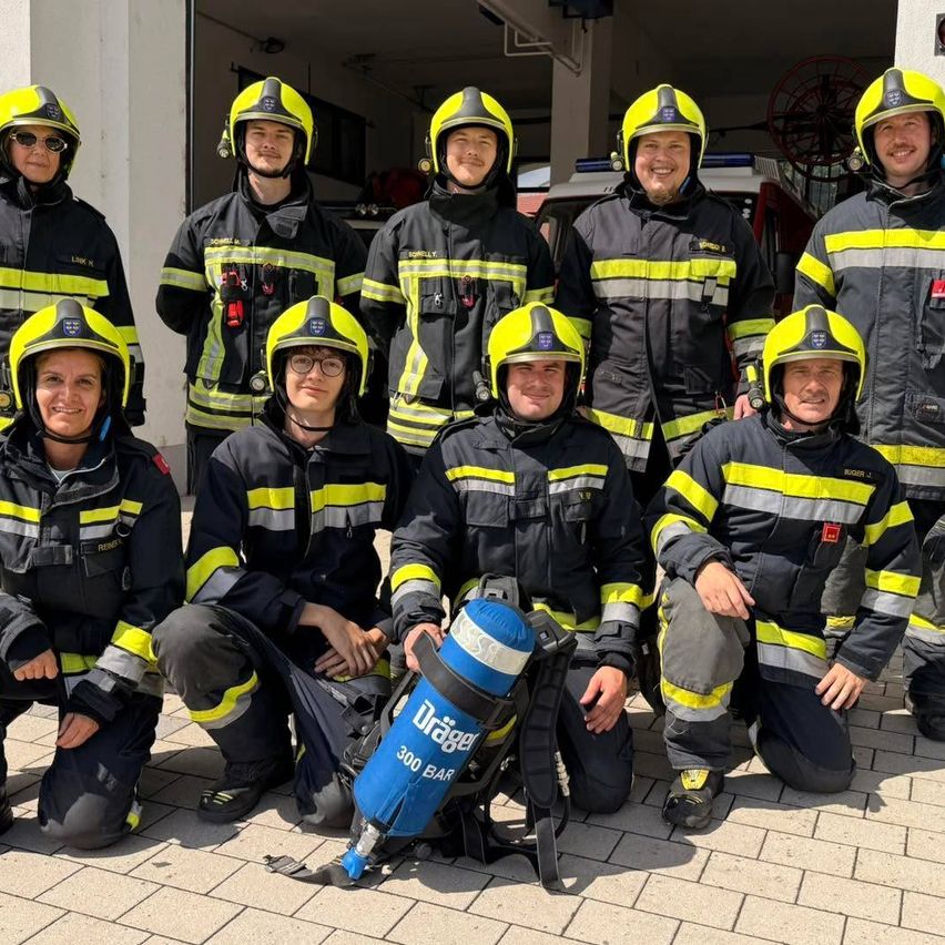 Eine Gruppe von Feuerwehrleuten in voller Uniform, lächelnd und posierend für ein Foto, wobei einer einen blauen Sauerstofftank hält.