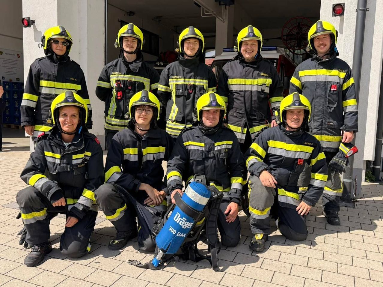 Eine Gruppe von Feuerwehrleuten in voller Uniform, lächelnd und posierend für ein Foto, wobei einer einen blauen Sauerstofftank hält.