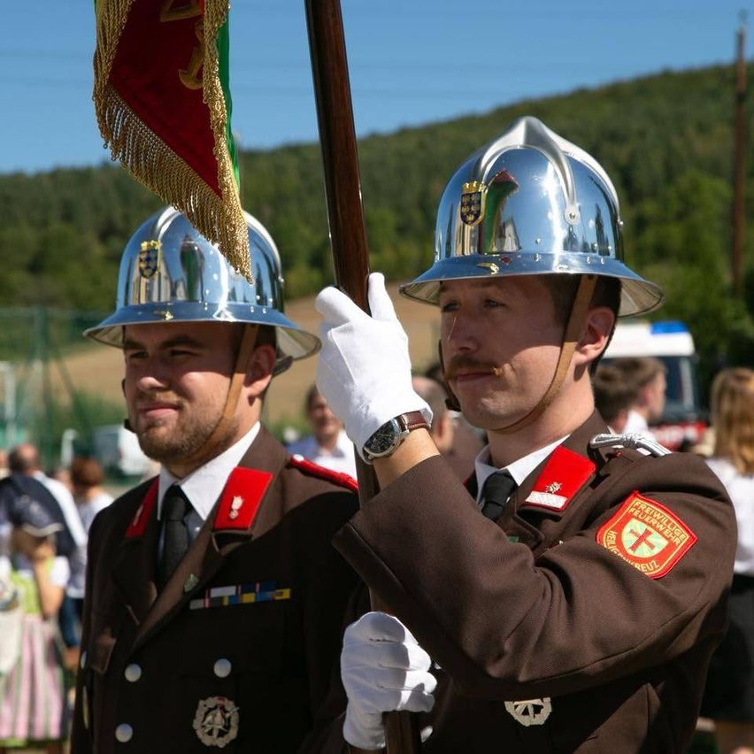 Zwei Feuerwehrleute in Uniform, einer hält eine Fahne, stehen vor einer Menge mit einem Berg im Hintergrund.