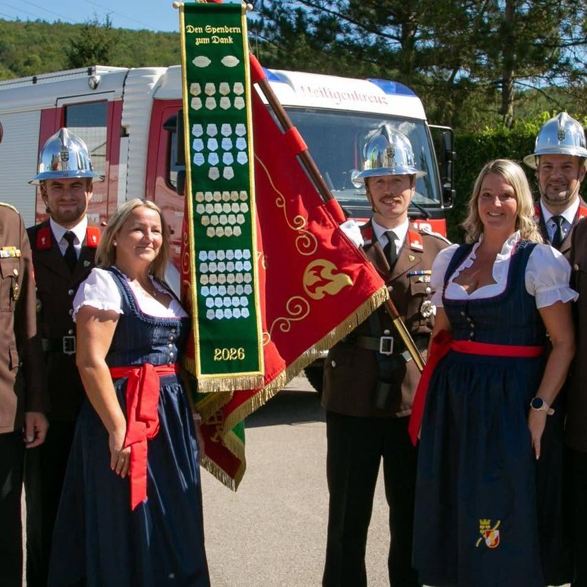 Eine Gruppe von Menschen, darunter zwei Frauen in traditioneller Kleidung, steht vor einem Feuerwehrwagen mit einer Fahne und einem Banner. Die Männer tragen Uniformen.