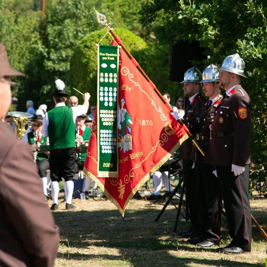 Eine Gruppe von Männern in traditionellen Uniformen und Hüten steht in einer Reihe und hält eine Flagge und ein Banner. Sie nehmen möglicherweise an einer Parade teil.