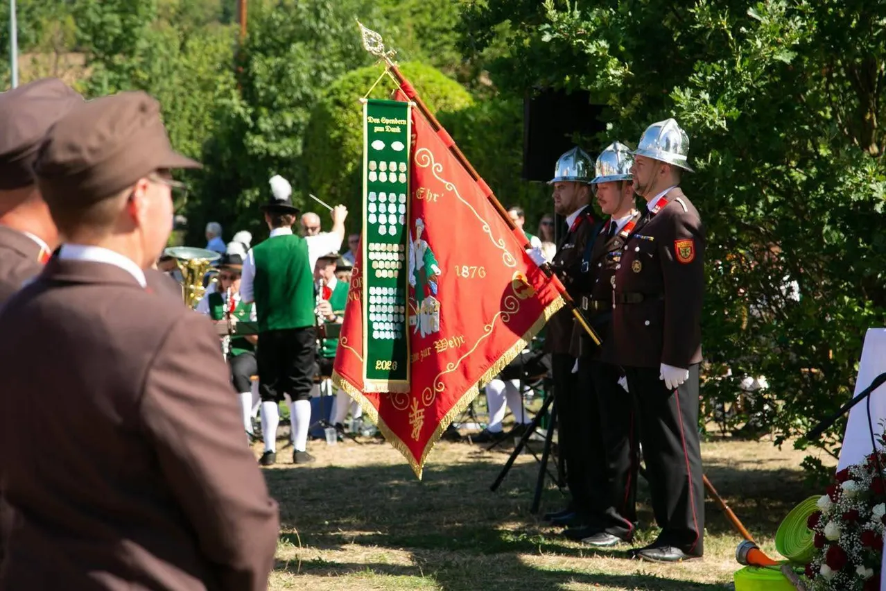 Eine Gruppe von Männern in traditionellen Uniformen und Hüten steht in einer Reihe und hält eine Flagge und ein Banner. Sie nehmen möglicherweise an einer Parade teil.