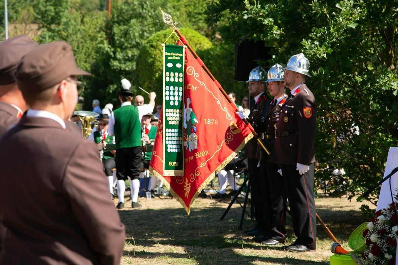 Eine Gruppe von Männern in traditionellen Uniformen und Hüten steht in einer Reihe und hält eine Flagge und ein Banner. Sie nehmen möglicherweise an einer Parade teil.