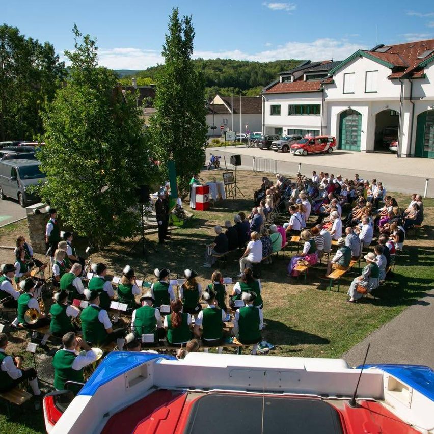 Eine Band spielt im Freien vor einem Publikum, mit einem großen Gebäude und geparkten Autos im Hintergrund.