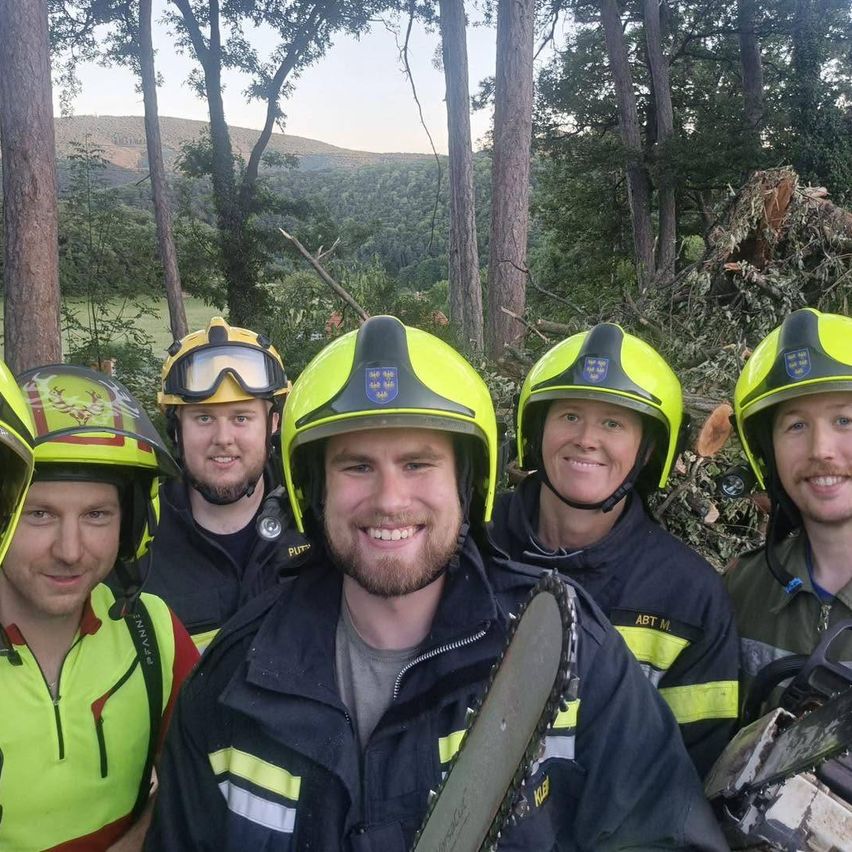 Eine Gruppe von Feuerwehrleuten in Schutzkleidung posiert für ein Foto in einem Wald mit Bäumen und Bergen im Hintergrund. Sie halten Kettensägen.