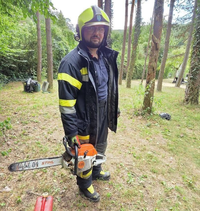 Ein Mann in Schutzkleidung steht mit einer Kettensäge in einem Wald und schaut in die Kamera.