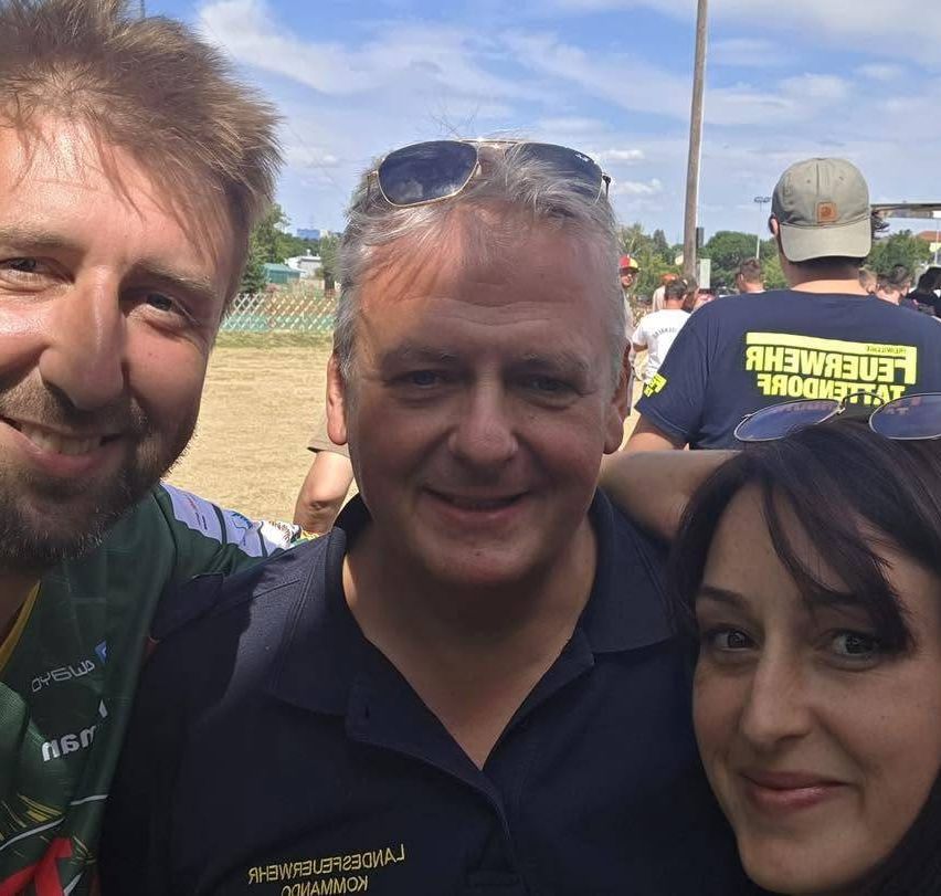 Drei Personen posieren für ein Foto im Freien. Der Mann in der Mitte trägt ein blaues Shirt. Ein Mann rechts trägt ein grünes Shirt mit einem gelben Logo.