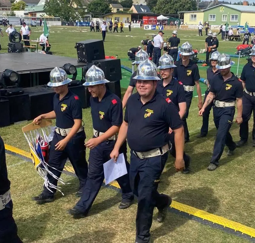 Eine Gruppe von Feuerwehrleuten in Uniform marschiert auf einem Grasfeld mit etwas Ausrüstung im Hintergrund.