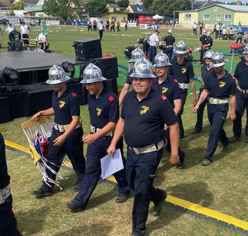 Eine Gruppe von Feuerwehrleuten in Uniform marschiert auf einem Grasfeld mit etwas Ausrüstung im Hintergrund.