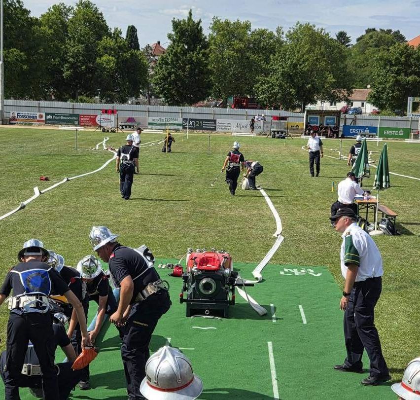 Eine Gruppe von Feuerwehrleuten übt ihre Fähigkeiten auf einem Feld mit grünen Markierungen. Sie tragen Helme, Schutzanzüge und tragen Ausrüstung. Eine Person biegt sich nach unten, während eine andere eine Schlange hält. Es gibt auch Leute, die stehen und beobachten.