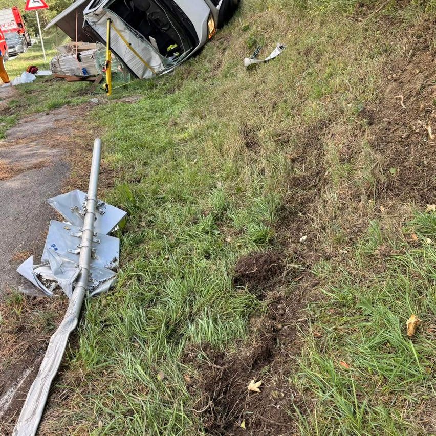 Ein umgestürztes Auto auf einem Grashang, mit einem umgestürzten Pfahl in der Nähe. Trümmer und ein Verkehrsschild sind im Hintergrund zu sehen.