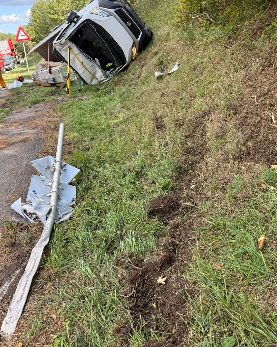 Ein umgestürztes Auto auf einem Grashang, mit einem umgestürzten Pfahl in der Nähe. Trümmer und ein Verkehrsschild sind im Hintergrund zu sehen.