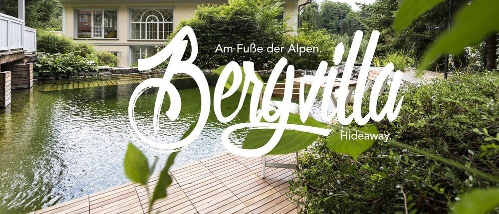 Eine ruhige Villa namens Bergvilla, am Fuße der Alpen gelegen, umgeben von üppiger Grünanlage und einem Teich, mit einer Holzterrasse und einem Liegestuhl.