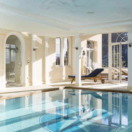 Ein Hallenbad mit blauem Wasser, Glastüren, Liegestühlen und dekorativen Fenstern, umgeben von einem weiß gestrichenen Raum.