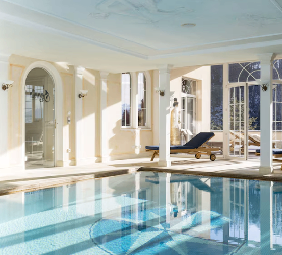 Ein Hallenbad mit blauem Wasser, Glastüren, Liegestühlen und dekorativen Fenstern, umgeben von einem weiß gestrichenen Raum.