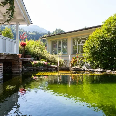 Eine ruhige Szene eines Hauses mit einer Holzbrücke über einem Teich, umgeben von üppiger Grünfläche und Blumen, mit Bergen im Hintergrund.