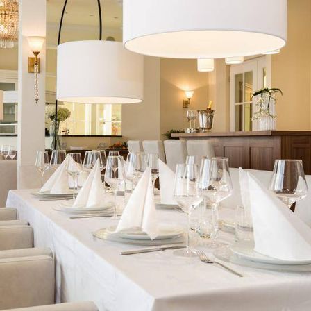 Ein eleganter Essbereich mit einem langen Tisch, der für ein formelles Event gedeckt ist. Jeder Stuhl ist mit weißem Tuch bespannt, und der Tisch hat gefaltete Servietten, Teller, Weingläser und Besteck.