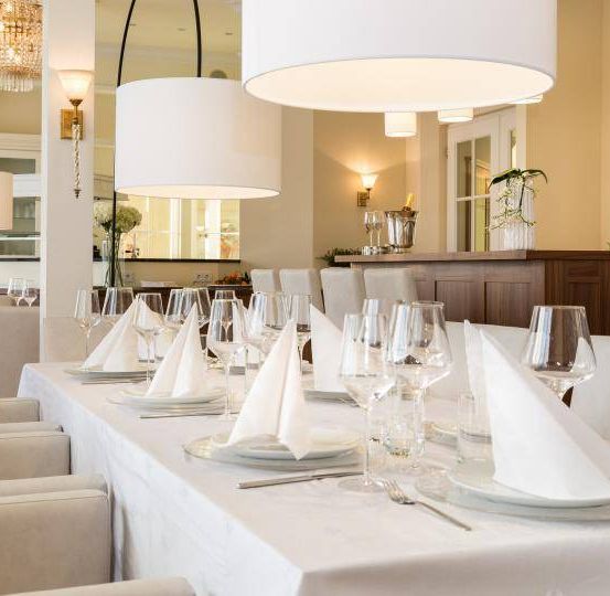 Ein eleganter Essbereich mit einem langen Tisch, der für ein formelles Event gedeckt ist. Jeder Stuhl ist mit weißem Tuch bespannt, und der Tisch hat gefaltete Servietten, Teller, Weingläser und Besteck.