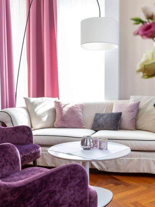 Ein Wohnzimmer mit einer weißen Couch, die mit verschiedenen Kissen verziert ist, einem weißen Tisch und einem rosa Stuhl. Eine weiße Lampe hängt von der Decke, und Blumen befinden sich rechts.