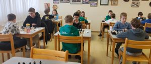 Eine Gruppe von Kindern spielt Schach in einem Klassenzimmer mit Holztischen und einem Holzboden, umgeben von verschiedenen gerahmten Bildern an der Wand.