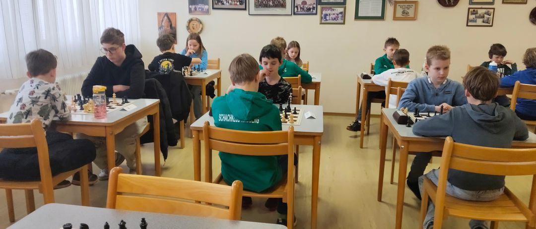 Eine Gruppe von Kindern spielt Schach in einem Klassenzimmer mit Holztischen und einem Holzboden, umgeben von verschiedenen gerahmten Bildern an der Wand.