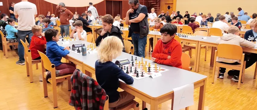 Eine Gruppe von Kindern spielt Schach in einer Halle. Sie sitzen an Tischen, mit einigen Erwachsenen, die um sie herum stehen.
