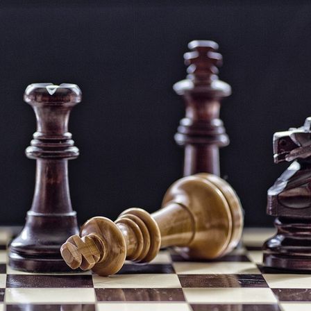 Ein Schachbrett zeigt ein gefallenes König-Stück mit einem Bischof und einem weiteren König, die noch stehen, auf einem schwarzen Hintergrund.
