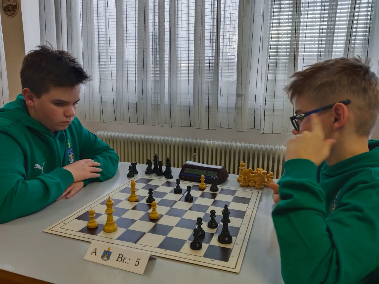 Zwei Jungen in grünen Pullovern spielen Schach auf einem Brett mit den Bezeichnungen A und Br. 5, mit einer digitalen Uhr in der Nähe. Sie sitzen vor einem Fenster mit Vorhängen.