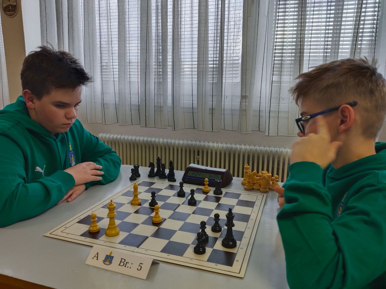Zwei Jungen in grünen Pullovern spielen Schach auf einem Brett mit den Bezeichnungen A und Br. 5, mit einer digitalen Uhr in der Nähe. Sie sitzen vor einem Fenster mit Vorhängen.