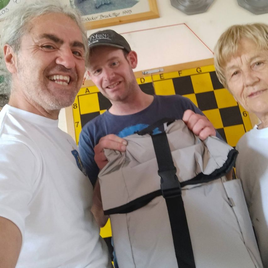 Drei Menschen lächeln und posieren für ein Foto. Der Mann links trägt ein weißes T-Shirt, der Mann in der Mitte trägt ein blaues T-Shirt und eine Kappe, und die Frau rechts trägt ein weißes T-Shirt. Der Mann in der Mitte hält eine graue Tasche.