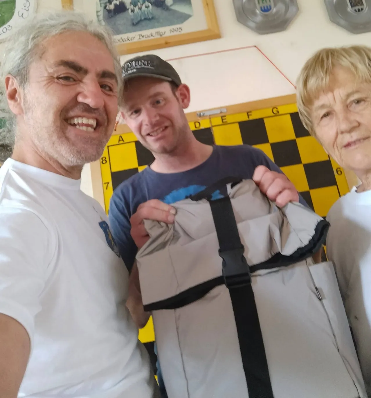 Drei Menschen lächeln und posieren für ein Foto. Der Mann links trägt ein weißes T-Shirt, der Mann in der Mitte trägt ein blaues T-Shirt und eine Kappe, und die Frau rechts trägt ein weißes T-Shirt. Der Mann in der Mitte hält eine graue Tasche.