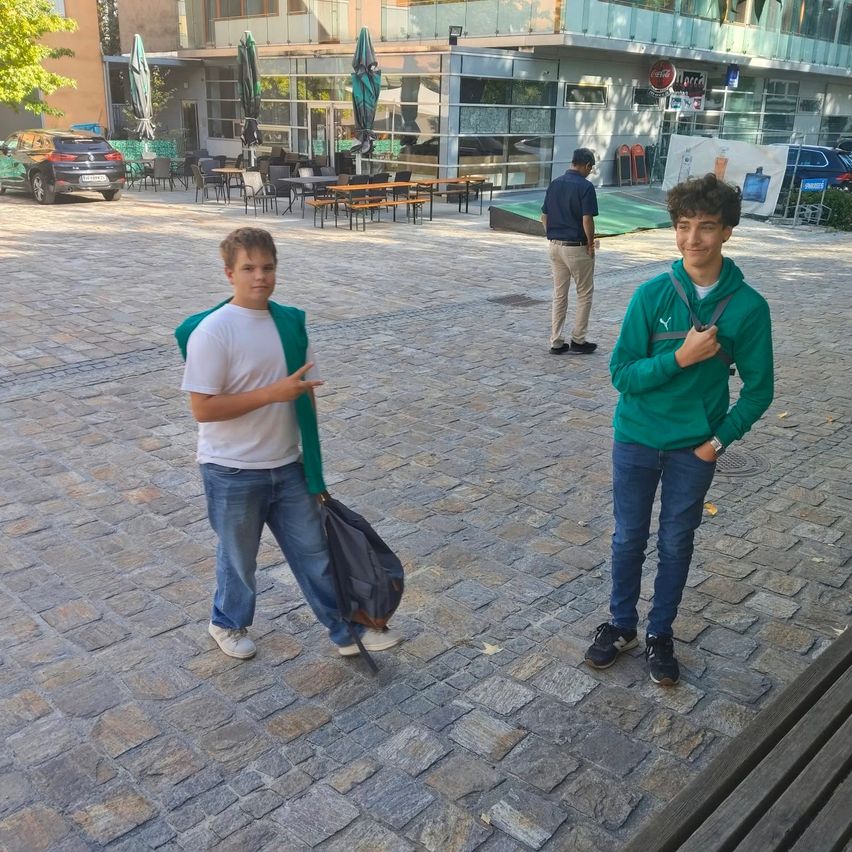 Zwei junge Männer stehen auf einem gepflasterten Platz, einer mit einer Tasche, der andere mit einer grünen Kapuze. Dahinter geht ein Mann, und es sind Tische und Stühle für ein Café im Freien aufgestellt.