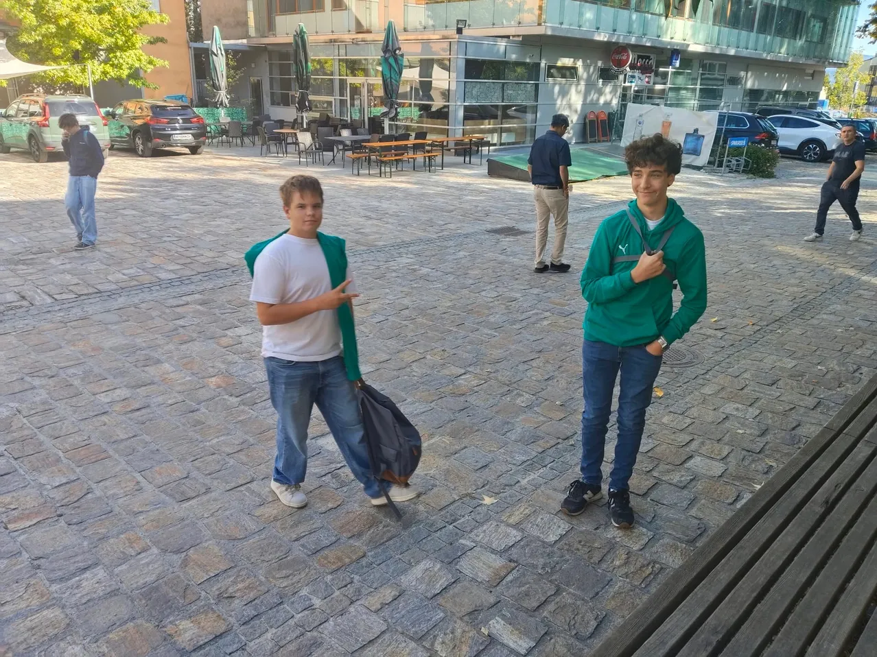 Zwei junge Männer stehen auf einem gepflasterten Platz, einer mit einer Tasche, der andere mit einer grünen Kapuze. Dahinter geht ein Mann, und es sind Tische und Stühle für ein Café im Freien aufgestellt.